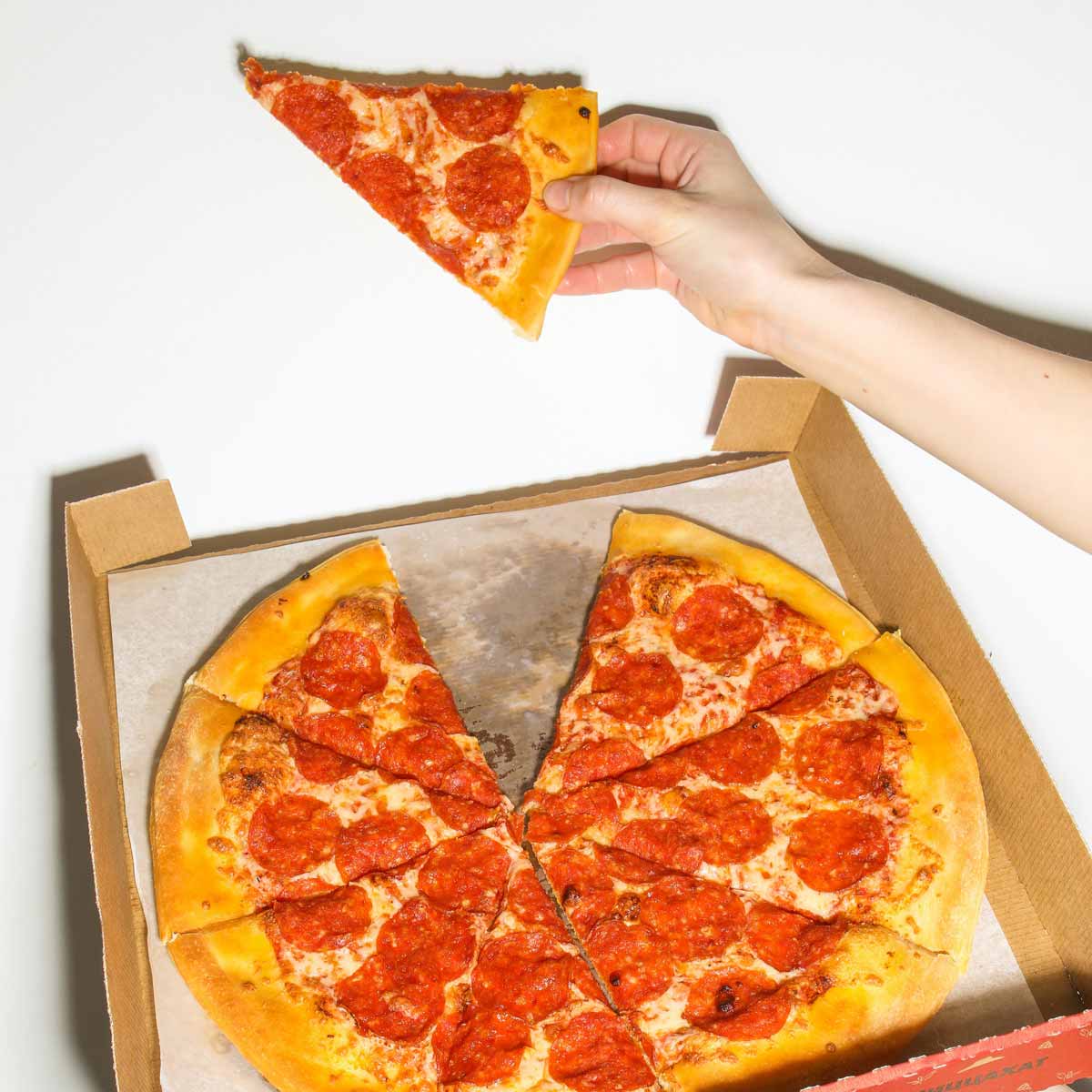 A hand holding a pepperoni pizza slice in front of a pizza in a pizza box